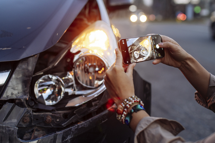 A person in a reflective vest takes a photo of a damaged car with a smartphone after a traffic accident. The image on the phone screen shows the front-end damage clearly. This scene is ideal for illustrating mobile insurance claims, accident documentation, roadside assistance, and vehicle emergencies.